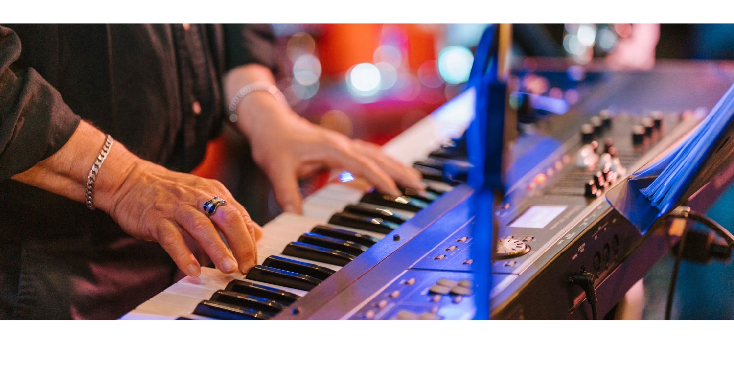 A close-up shows a musician's hands with a ring and bracelet playing a black and white electronic keyboard piano under vibrant, colorful stage lighting.